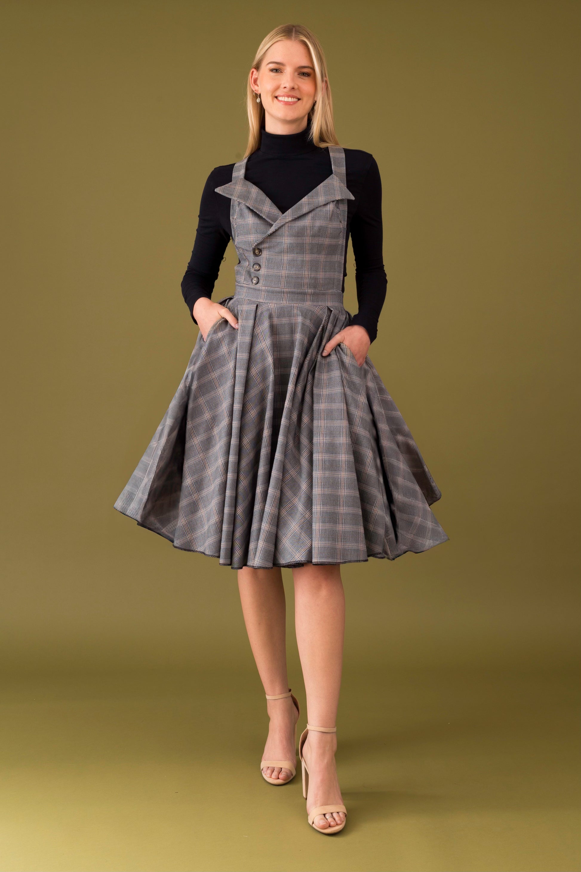 A person wearing a midi dress with turtleneck collar, made from checkered fabric, featuring a pinafore style with lapels and straps, and pockets on the side.