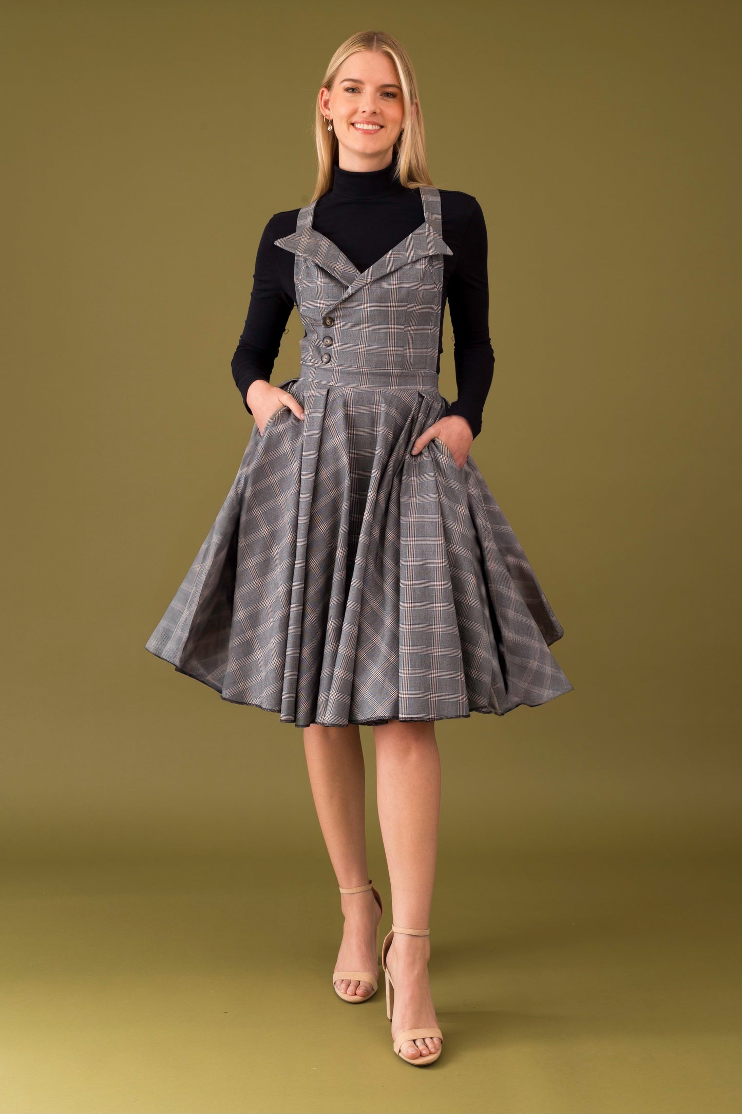 A person wearing a midi dress with turtleneck collar, made from checkered fabric, featuring a pinafore style with lapels and straps, and pockets on the side.